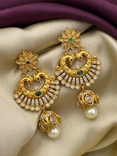 Exquisite chandbali earrings, finished in a matte gold polish, features a captivating floral stud highlighted by a vibrant emerald-green stone at its center, leading to an elegantly curved crescent adorned with Peacock design work. The crescent is embellished with sparkling white stones that mimic the brilliance of diamonds, adding a touch of luxury. Dangling gracefully, a lustrous pearl drop ensconced in a golden Jhumka type completes the ensemble, offering a classic yet contemporary appeal. Golden Jhumka, Emerald Green Stone, Traditional Indian Jewellery, Chandbali Earrings, Floral Studs, White Stones, Bollywood Style, Peacock Design, Cz Jewelry