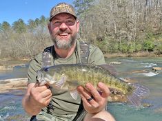 a man holding a fish while sitting on a rock in the water with trees behind him