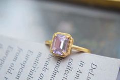 This ring features a central rectangular gemstone with a soft pink hue, cut in an emerald style with step facets that create a classic and elegant look. The stone is set in a gold band with four prongs securing it at each corner, and the setting is bordered by a decorative, textured frame that adds to the ring's vintage appeal. The overall design combines simplicity and sophistication, making it a charming and timeless piece. Details Isabella Rectangular Crystal Vintage Ring Adjustable opening Gold band Rare, antique design Reyis Jewelry Isabella Collection Formal Pink Rectangular Ring, Gold Rectangular Crystal Ring, Elegant Gold Rectangular Crystal Ring, Formal Pink Rings With Rectangular Stone, Elegant Ring With Faceted Rectangular Stone, Elegant Crystal Ring With Rectangular Stone, Elegant Crystal Ring With Rectangular Gemstone, Elegant Pink Rings With Rectangular Stone, Classic Pink Square Cut Ring