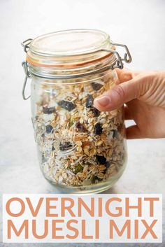 a hand holding a jar filled with oatmeal muesli mix on top of a table