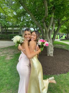 two women hugging each other in front of a tree with flowers on the ground and grass