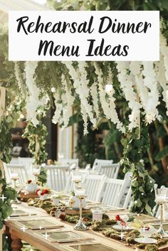 an outdoor dinner table with white chairs and flowers hanging from the ceiling, surrounded by greenery