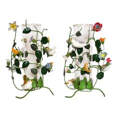 two glass vases with flowers and leaves in them on metal stands against a white background