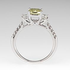 This beautiful yellowish green sapphire ring is centered with one (1) cut-cornered square mixed cut natural sapphire weighing 1.33 carats and set into a four-prong head. The sapphire is flanked to each side by one (1), channel/prong set, square modified brilliant cut diamond. The top face of the shank is accented with five (5), prong set, round brilliant cut diamonds. The ring measures 6.6mm at the top, rises 7.3mm above the finger, tapering to 1.6mm wide and 1.3mm thick at the base of the shank. The ring is currently size 5.5. Yellow Emerald Cut Diamond Ring, Gia Certified Square Cut Sapphire Diamond Ring, Square Cut Sapphire Ring With Diamond Prong Setting, Square Cut Sapphire Ring With Diamond, Gia Certified Asscher Cut Sapphire Ring, Green Sapphire Ring, Yellowish Green, Three Stone Engagement Ring, Stone Engagement Ring