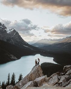 two people sitting on top of a large rock next to a body of water with mountains in the background