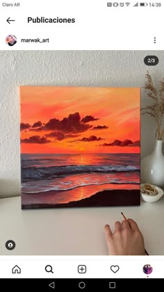 a person is holding a pen and looking at a painting on the table with an orange sunset in the background