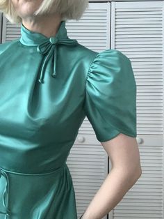 "This stunning tetal satin dress by House of Bianchi dates to the '70s and features hidden back zip closure, attached waist tie, split tulip puff sleeve, off-center Mandarin collar with bow, and pleating at neckline/bust.  Materials:  Fabric unknown but appears to be a heavy satin polyester. Unlined.  Measurements:  Chest: 34\" Waist: 26\" Waist length: 15\" Shoulder: 15\" Total length: 44\" Shown on size S at 5'3\" Measurements taken laid flat  Please be certain before you buy this item because I do not accept returns. Please take care to check the measurements before you purchase as there are no returns for poor fit. We carefully measure each item, and measurements are given seam-to-seam, with minimal stretching. Depending on how much you stretch the fabric, the measurements differ sligh Fitted Silk Vintage Dress, Fitted Silk Vintage Dress For Vintage Fashion, Fitted Vintage Satin Dress, Retro Fitted Satin Dress, Fitted Retro Satin Dresses, Vintage Fitted Satin Dress, Retro Fitted Silk Dress, Elegant Fitted Satin Vintage Dress, Mandarin Collar Dress