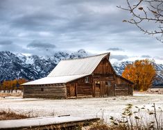 an old barn sits in the middle of a field with mountains in the background and snow on the ground