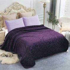 a bed with purple comforter and pillows in a room next to a lamp on a table