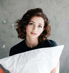 a woman sitting on top of a bed holding a white pillow and looking at the camera