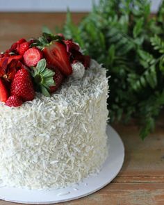 a white cake topped with strawberries and coconut on top of a wooden table next to greenery