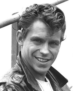 a black and white photo of a man with curly hair smiling in front of a pole