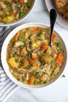 two bowls of chicken and vegetable soup on a white table with bread in the background