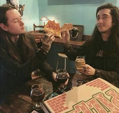 two men are sitting at a table eating pizza and drinking beer while one man holds up a slice of pizza