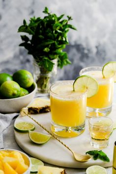 two glasses filled with orange juice and limes on a white platter next to bowls of fruit