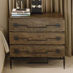 a nightstand with a clock and books on it in front of a curtained window