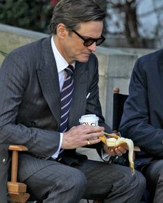 a man in a suit and tie eating a banana while sitting on a wooden chair