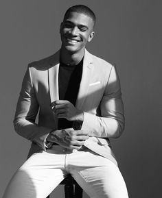 a black and white photo of a smiling man in a suit sitting on a stool