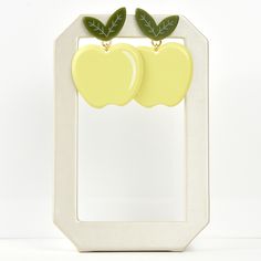 two yellow apples with green leaves hang from the back of a white frame, against a white background