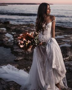 a woman in a wedding dress standing on rocks near the ocean