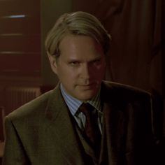 a man with blonde hair wearing a suit and tie in a dimly lit room, looking at the camera