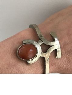 "I wish I could tell you more about the origins of this cuff or the metal and stone that it's made up of. But, what I can tell you that it is one of the most unique pieces of jewelry I have ever seen. The metal is silver tone and hard, possibly nickel. The stone is smooth and brown, probably agate. The poetry of this bracelet lies in its beautiful lines and handmade appearance. The face of this cuff is 1 3/4\" long and 1\" at its widest. The stone is oval shaped and bezel set. There are makers m Adjustable Oval Modern Cuff Bracelet, Modernist Metal Cuff Bracelet As Gift, Modernist Metal Cuff Bracelet Gift, Unique Cabochon Cuff Bangle Bracelet, Modernist Handmade Adjustable Cuff Bracelet, Modernist Adjustable Cuff Bracelet As Gift, Handmade Adjustable Modernist Cuff Bracelet, Unique Oval Cuff Bracelet For Gift, Vintage Oval Cuff Bracelet With Polished Finish