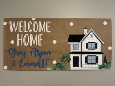 a welcome home sign with a house and trees painted on the front wall in brown, white and blue