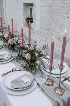 the table is set with white plates and silverware, pink candles and greenery