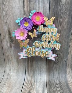 a birthday cake topper with flowers and butterflies on it's side, sitting on a wooden surface