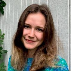 a woman with long brown hair wearing a blue shirt and smiling at the camera while standing in front of a white wall