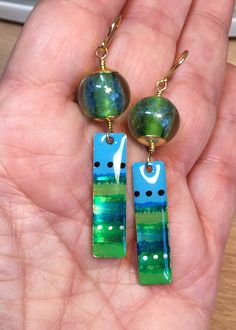 Springy Greens These earrings are created from artisan made glass beads. The round beads have a core of spring green, with a net-like coat of blue over them. They are encased with a thick coat of clear glass which magnifies the color and texture. Below them dangle artisan brass charms, 1 1/8 inch long and 3/8 inch wide. They have been painted with irregular stripes in a variety of blues and greens, both flat and sparkly, with dots of black and gold, then sealed with a thick coat of resin. I have Artsy Blue Beaded Dangle Earrings, Blue Artsy Beaded Dangle Earrings, Handmade Artsy Blue Beaded Earrings, Artsy Blue Beaded Earrings, Green Artisan Enamel Earrings, Artisan Green Enamel Earrings, Green Hand Painted Drop Earrings, Green Artisan Round Bead Earrings, Unique Green Round Beads Earrings