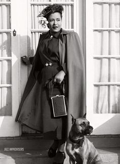 a woman standing next to a dog in front of a door wearing a cape and holding a purse