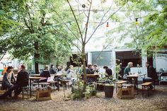 a group of people sitting at picnic tables in the shade of trees, with food trucks behind them