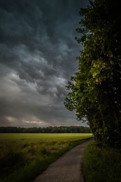 an empty road in the middle of a grassy field under a dark sky with storm clouds