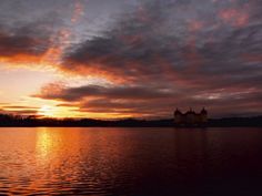 the sun is setting over a lake with some buildings in the distance and clouds above it