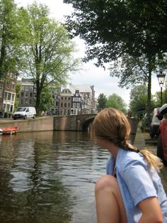 a woman sitting on the edge of a body of water next to trees and buildings