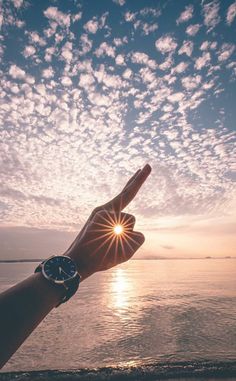 a person's hand pointing at the sun with clouds in the sky above them