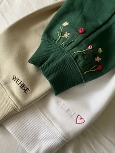 two sweaters with embroidered flowers on them are laying next to each other and one has a heart