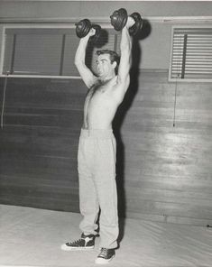 a shirtless man lifting two dumbbells over his head in an old black and white photo