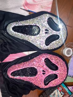 two masks are sitting on the floor next to some books and pens, with one mask covered in glitter