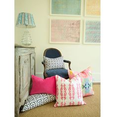 a chair with many pillows on it in front of some pictures and a table lamp