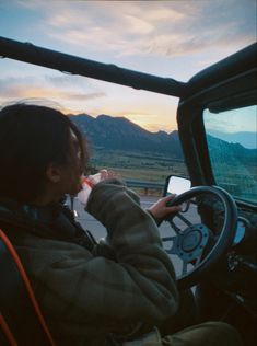 a person driving a truck with mountains in the background at sunset or sunrise, while wearing a hoodie and holding a steering wheel