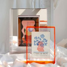 two framed pictures sitting next to each other on a white sheet covered tablecloth with an orange frame