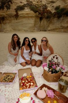 three women are sitting on the beach with pizzas and drinks in front of them