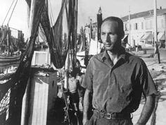 a man standing on the deck of a boat in front of other boats and buildings