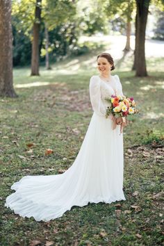 a woman in a white wedding dress holding a bouquet and standing on grass with trees behind her