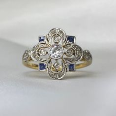 This item has just been reserved by another customer. Please contact us to be notified if it becomes available.  This handcrafted ring from the Edwardian period features an old European cut diamond weighing roughly 0.17 carats, with J color and SI2 clarity, at its center. Surrounding the center stone are four rose cut diamonds set in bezel, and a halo of floral motif rose-cut diamonds. Additionally, the design is accented by four antique French cut sapphires with a total weight of about 0.12 car Heirloom Diamond White Diamond Ring Hallmarked, Diamond White Sapphire Heirloom Ring For Anniversary, Elegant Gold Sapphire Ring With Single Cut Diamonds, Heirloom Sapphire Ring With Intricate Design For Anniversary, Heirloom Style Sapphire Ring With Diamond Cut, Hallmarked Sapphire Ring With Diamond In Round Cut, Antique Sapphire Diamond Ring For Wedding, Heirloom Style Hallmarked Diamond Cluster Ring, Heirloom Hallmarked Diamond Ring