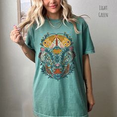 👀Images are an accurate depiction of what you'll receive upon purchase ♂️ All Shirts are Unisex Sizing 👣 Printing is "Direct To Garment"  (Ink printed). Step into a world of enchantment with our Mystical Moth T-shirt. This captivating piece of wearable art combines the timeless allure of folk art with the ethereal vibes of fairycore aesthetics. The graphic on this tee features a majestic moth, its wings adorned with intricate patterns reminiscent of ancient folk art. Surrounding the moth, a ce Green Casual Shirt With Vintage Print, Casual Green Shirt With Vintage Print, Green Bohemian Top With Graphic Print, Green Vintage Print Short Sleeve Top, Artistic Green Crew Neck T-shirt, Green Vintage Print Tops For Summer, Green Tops With Vintage Print For Summer, Spring Vintage Print Top With Short Sleeves, Multicolor Tops With Front Print For Summer