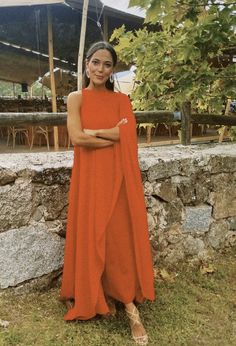 a woman wearing an orange dress standing in front of a stone wall with her arms crossed