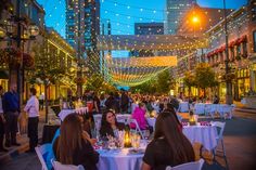 people sitting at tables in the middle of a city street with lights strung over it